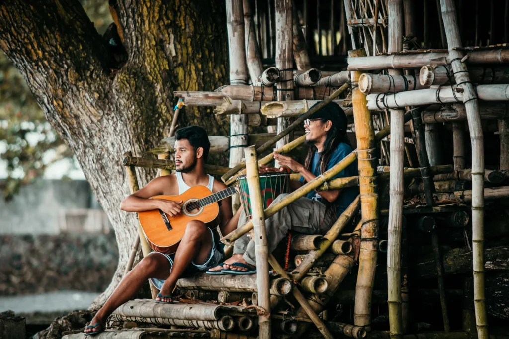 Two men relaxing and playing guitar and drum on a bamboo structure outdoors. Photo by James Ampong Quilario